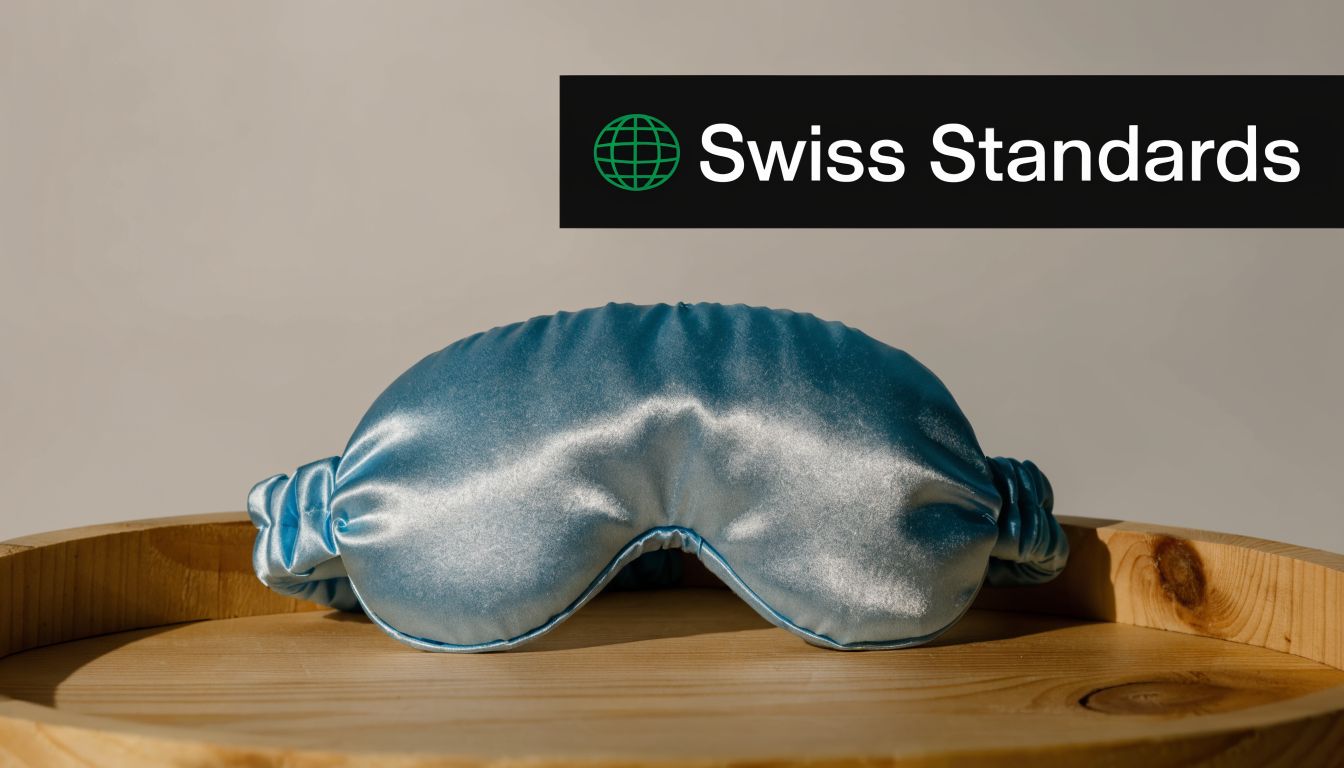 A light blue silky sleep mask resting on a wooden tray with a Swiss Standards logo overlay.
