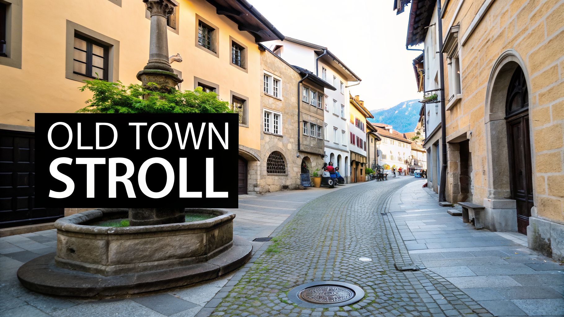 A charming old town street with historic buildings, a stone fountain, cobblestone road, and distant mountains.
